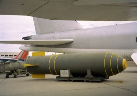 1977 Ford F-100 - A large bomb rests on a stand in front of an airplane, with another bomb visible on a cart to the left.