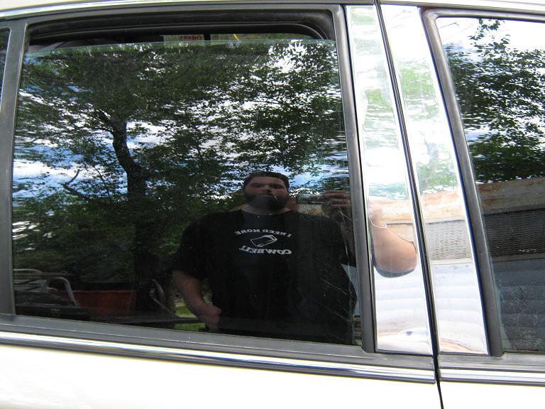 Reflection of a man in the side window of a 2000 Mercury Grand Marquis, showing chrome trim and a tree in the background