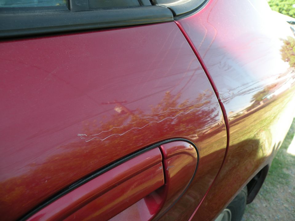 Close-up of long white scratch on rear door of dark red sedan, tree reflections in paint