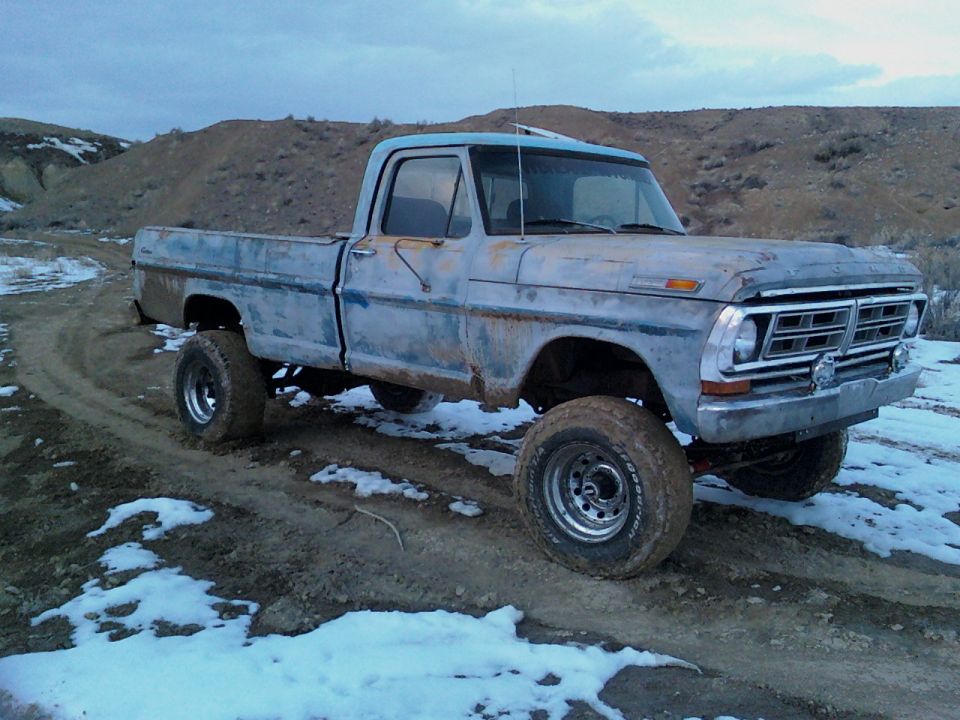 Blue 1970 Ford F-100 pickup truck with lifted suspension and off-road tires, parked on a dirt road with patches of snow