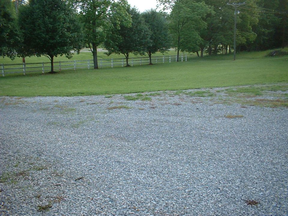Gravel driveway in foreground, green lawn with trees and white fence in background