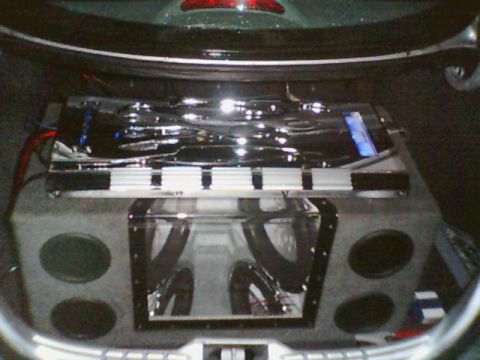 1998 Ford Taurus - car-audio install in a trunk: a chrome-finished amplifier sits atop a subwoofer box with a clear acrylic display window.