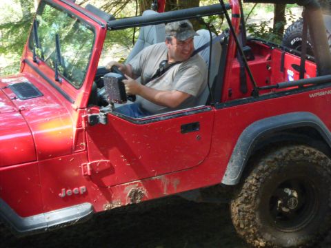 2005 Jeep Wrangler - A man drives a red Jeep Wrangler off-road, with mud splattered on its side.