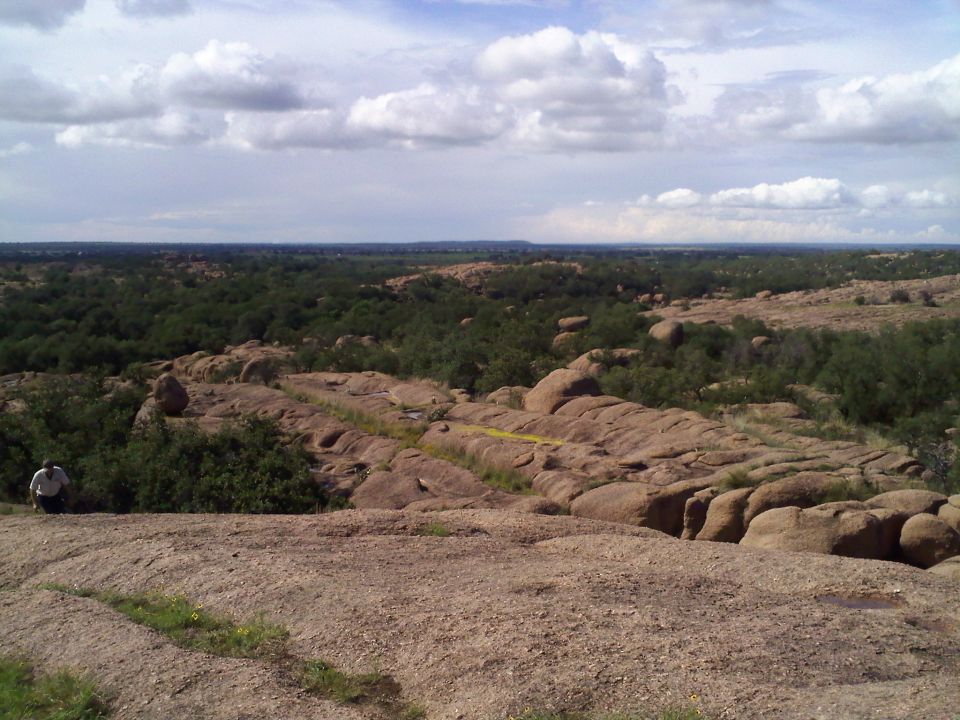 Rocky terrain with scattered trees under a cloudy sky. A person is visible on the left slope. Watermark reads SUPERMOTORS.NET
