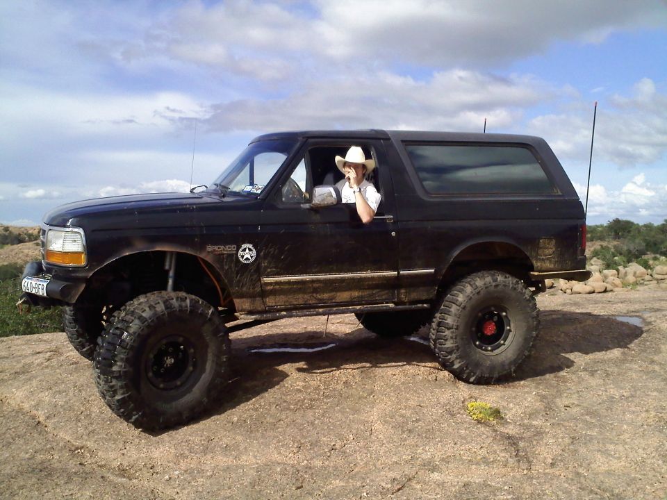 1995 Ford Bronco with lifted suspension and large off-road tires parked on rocky terrain, driver wearing a cowboy hat