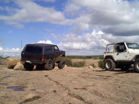1995 Ford Bronco - Two off-road vehicles, a black Ford Bronco and a white Jeep, are parked on a rocky landscape with a partly cloudy sky above.