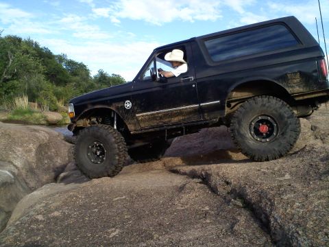 1995 Ford Bronco - A black 1995 Ford Bronco navigates a rocky off-road path under a blue sky with scattered clouds.
