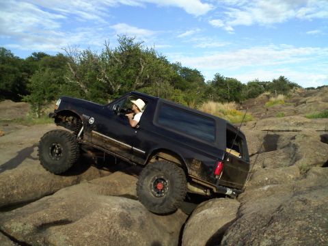 1995 Ford Bronco - A black Ford Bronco is shown off-roading over large rocks under a partly cloudy sky.