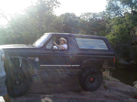 1995 Ford Bronco - A 1995 Ford Bronco is shown off-roading on a rocky terrain with trees in the background.