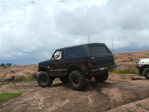 1995 Ford Bronco - A black 1995 Ford Bronco navigates a rocky landscape, with another vehicle visible in the background.