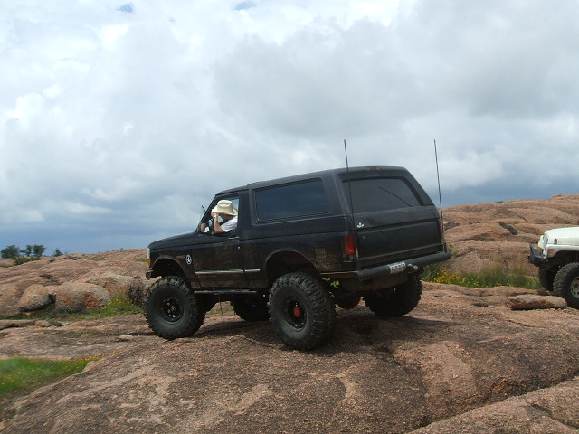 Black 1995 Ford Bronco with large tires on rocky terrain, driver wearing a hat. Another white vehicle is partially visible to the right