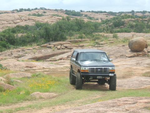 1995 Ford Bronco - A lifted 1995 Ford Bronco navigates a , rocky landscape under a cloudy sky.