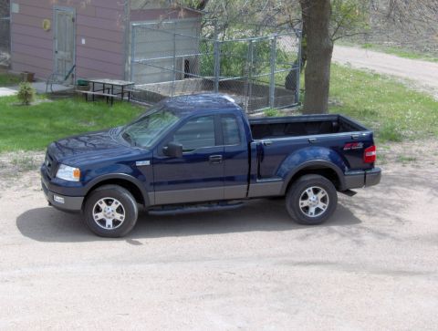 2005 Ford F-150 - A blue Ford F-150 pickup truck is parked on a dirt road near a building and a tree.