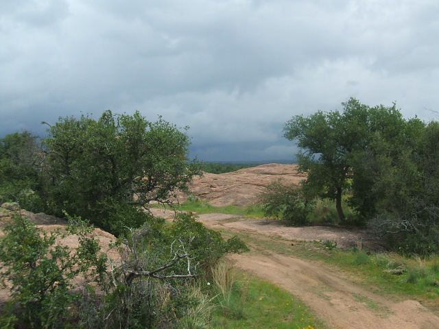 Dirt road winds through a rocky, tree-filled landscape under a cloudy sky