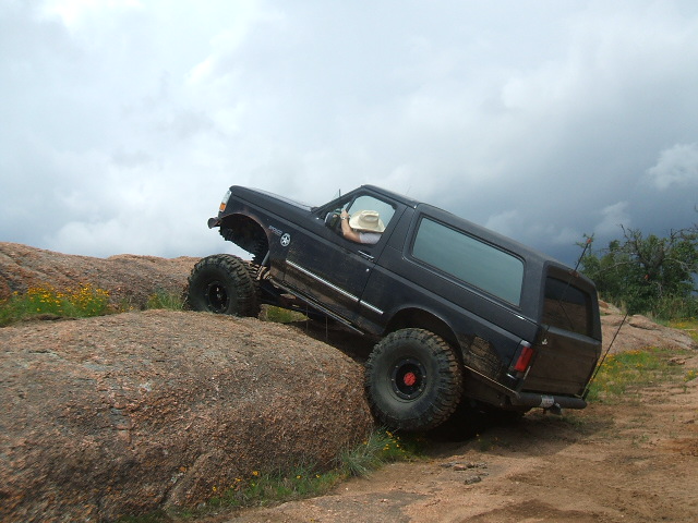 Black 1995 Ford Bronco with large tires climbing a rocky incline under a cloudy sky