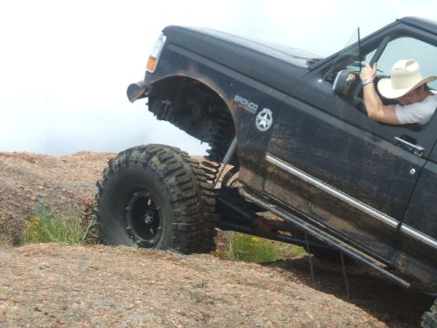 1995 Ford Bronco - A black 1995 Ford Bronco navigates rocky terrain, its large tires gripping the surface as the driver, wearing a cowboy hat, steers.