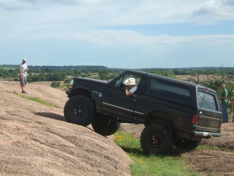 1995 Ford Bronco - A black 1995 Ford Bronco is shown off-roading on a large rock formation under a partly cloudy sky.