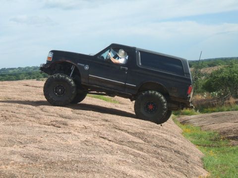 1995 Ford Bronco - A modified black Ford Bronco navigates a steep, rocky incline under a partly cloudy sky.