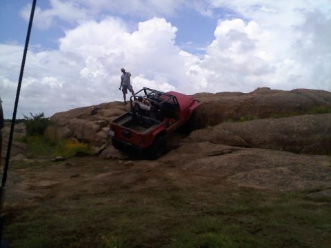1995 Ford Bronco - A red off-road vehicle navigates a challenging rocky terrain with a person standing on a boulder nearby.