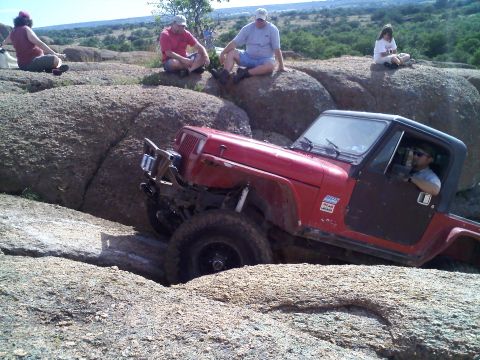1995 Ford Bronco - A red Jeep Wrangler is shown off-roading through a rocky landscape with several people watching.