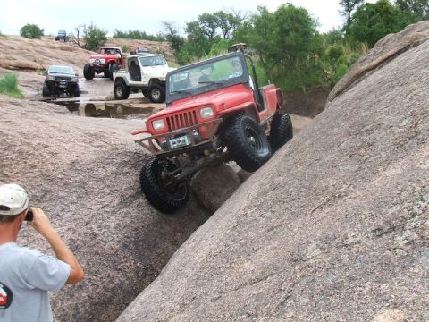 1995 Ford Bronco - Several off-road vehicles navigate a challenging rocky terrain, with a red Jeep Wrangler prominently ascending a steep boulder.
