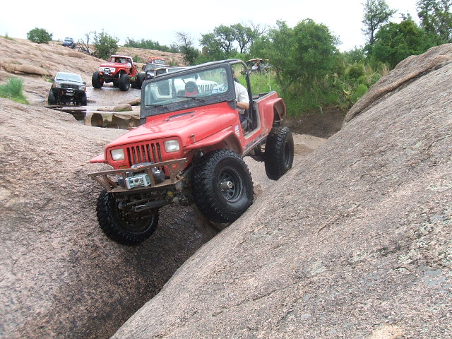 Red Jeep Wrangler climbing a rocky incline with other off-road vehicles in the background