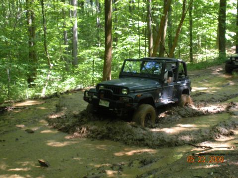 1998 Chevrolet Blazer - A dark-colored Jeep Wrangler navigates a muddy trail through a forest, with another vehicle visible in the background.