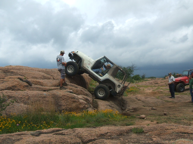 White Jeep Wrangler climbing a rocky incline with a person standing nearby; red Jeep visible in background