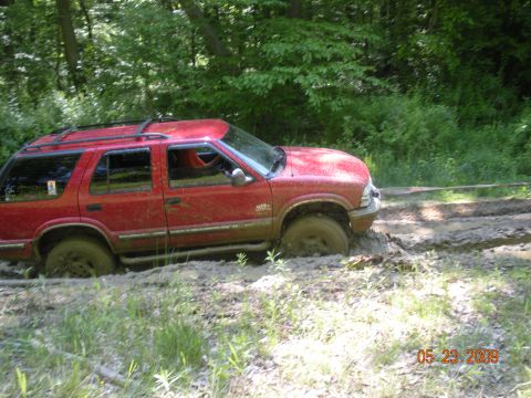1998 Chevrolet Blazer - A red 1998 Chevrolet Blazer is stuck in the mud on a wooded trail, with trees and greenery in the background.