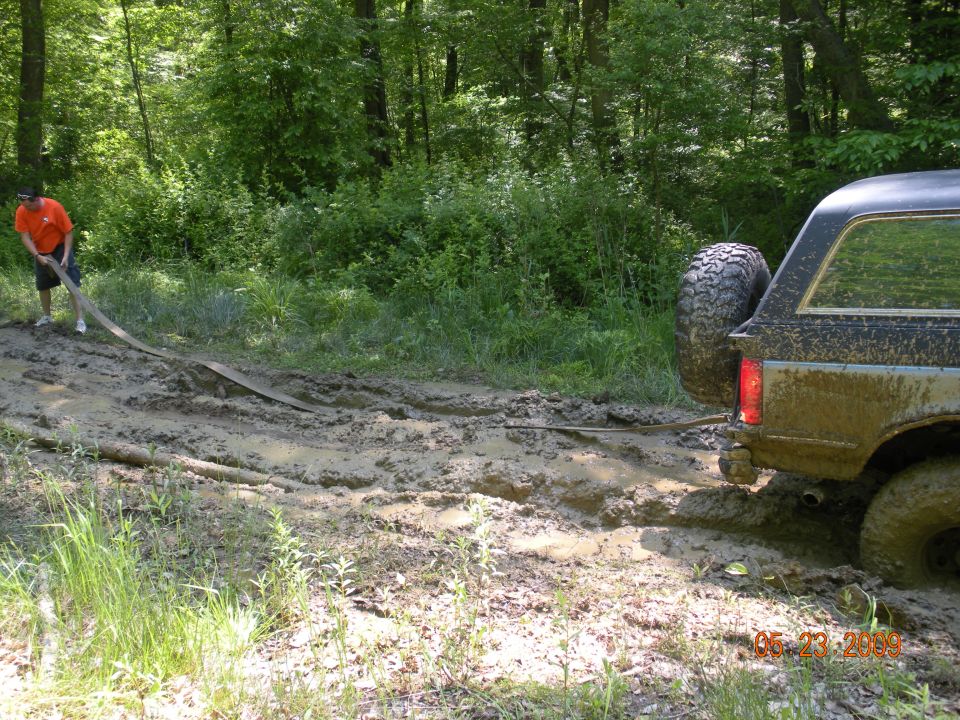 guy pulling a strap attached to a muddy SUV stuck in a dirt path in the woods