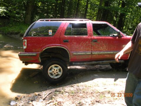 1998 Chevrolet Blazer - A muddy red 1998 Chevrolet Blazer navigates through a stream in a wooded area.