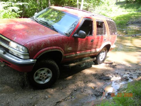 1998 Chevrolet Blazer - This red SUV appears to be stuck in a muddy, wooded area, with water and mud visible around its tires.