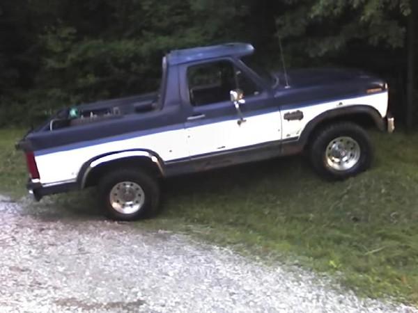 Dark blue and white Ford Bronco with rear top removed, side view on grass by a gravel drive