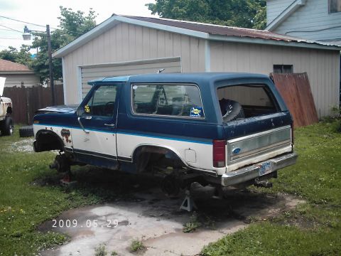 1986 Ford Bronco - Ford Bronco on jack stands in a yard, missing its wheels and tires.