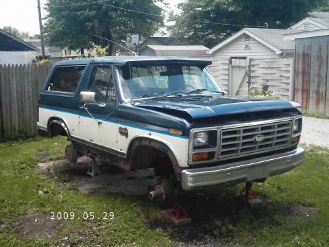 1986 Ford Bronco - A two-tone Ford Bronco sits on jack stands in a yard, missing its wheels and showing damage to the hood.