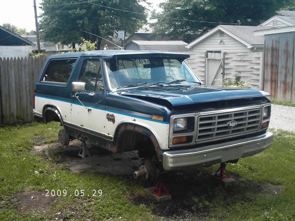 Dark blue and white Ford Bronco on jack stands, missing wheels, with a dented hood