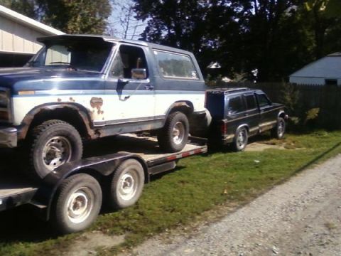 1994 GMC Jimmy - A blue and white SUV is loaded onto a trailer, with a black SUV parked nearby on a grassy area.