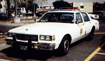 1991 Ford LTD Crown Victoria - A white police sedan with emergency lights on the roof is parked in a lot.