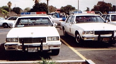 1991 Ford LTD Crown Victoria - Two white police cruisers, identified as 1991 Ford Crown Victoria LTDs, are parked in a lot.