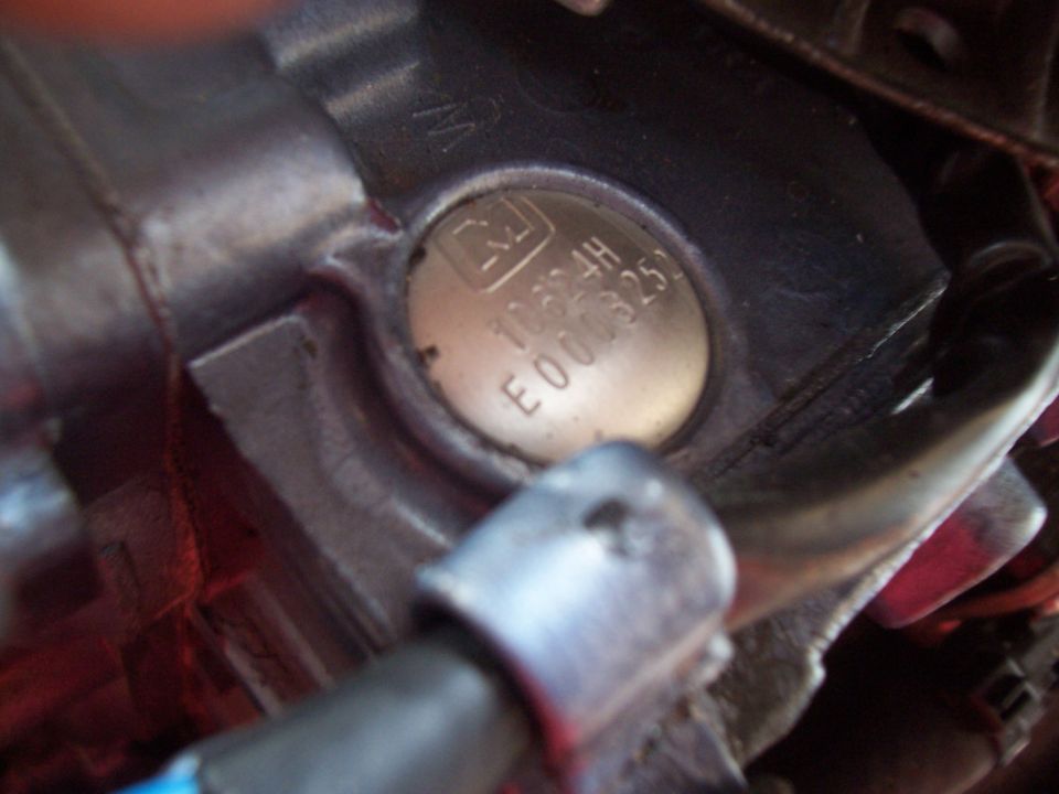 Close-up of a metal engine component with a circular stamped identification plate showing text and numbers