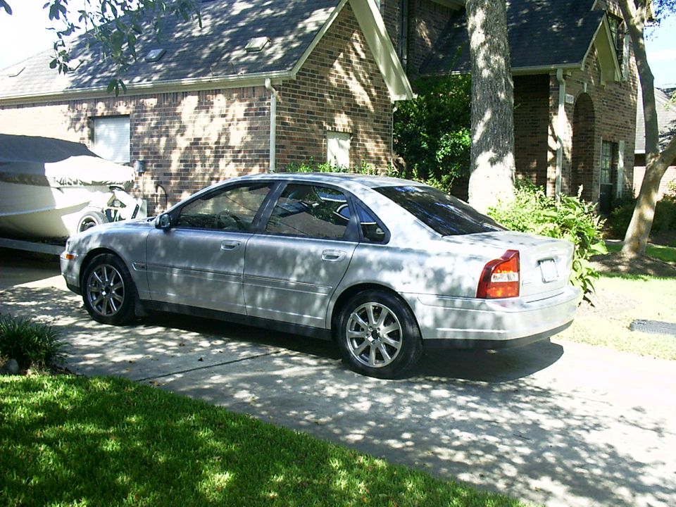 Silver Volvo S80 sedan in a driveway in front of a brick house, rear-quarter view