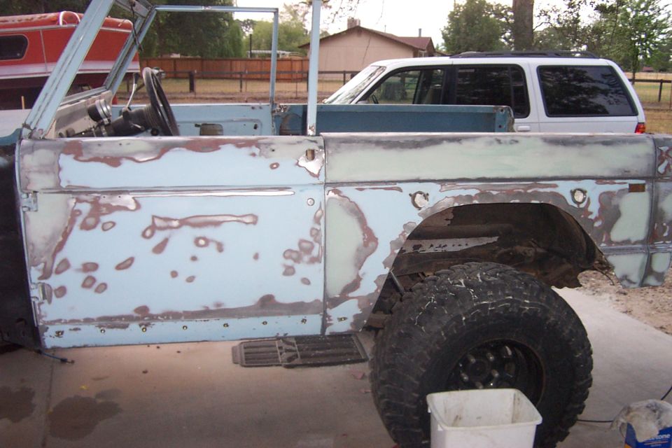 Side view of a light blue Ford Bronco body, showing primer and rust spots. A white SUV is parked behind it