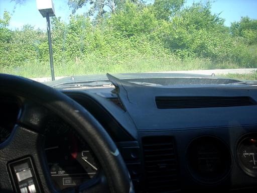 Black vehicle interior dashboard with cracked top and gauges, greenery visible through windshield