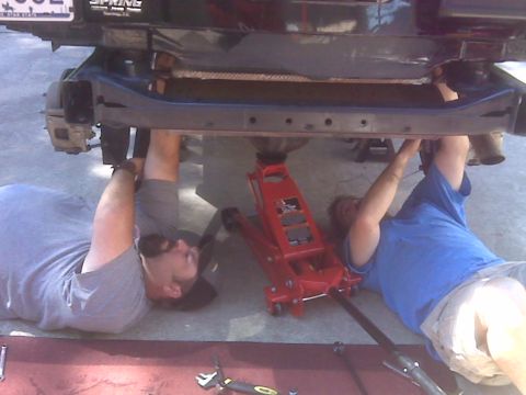 2008 Jeep Wrangler - Two people are working on the underside of a 2008 Jeep Wrangler lifted by a floor jack.