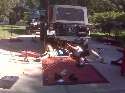 2008 Jeep Wrangler - Two people are working on the underside of a Jeep Wrangler in a driveway, with another Jeep parked behind it.