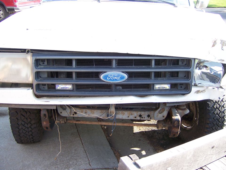 Front view of a damaged white Ford Bronco with a black grille and Ford logo. The bumper is bent and the headlight is broken