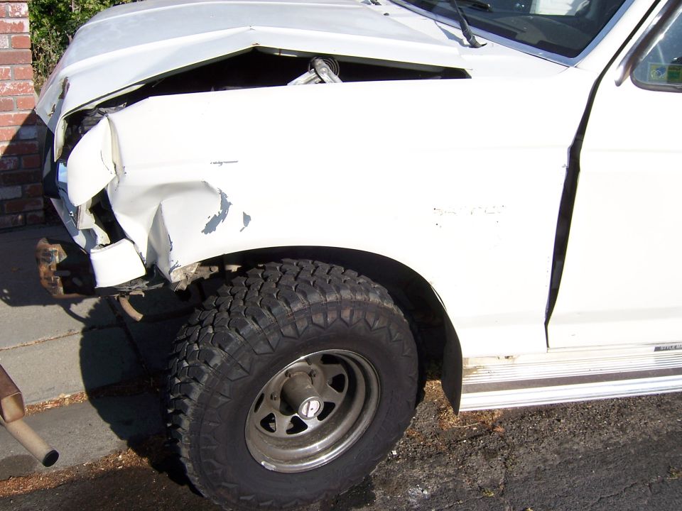 Front corner of white SUV with heavy front-end damage. Tire and fender are visible