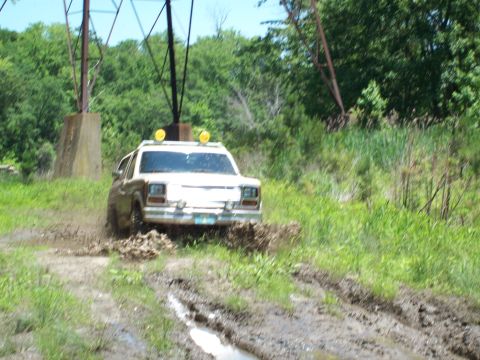 1986 Ford Bronco - This white SUV navigates a muddy trail, kicking up dirt and water.