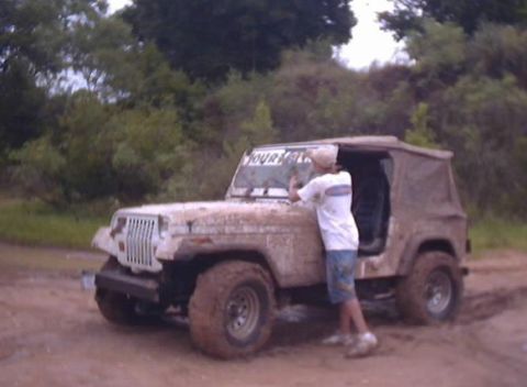1989 Ford Bronco - A person cleans the windshield of a muddy Jeep Wrangler off-roading in a wooded area.