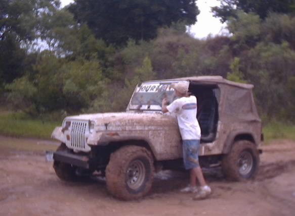 Mud-covered Jeep Wrangler with a person cleaning the windshield. Trees and dirt road in background
